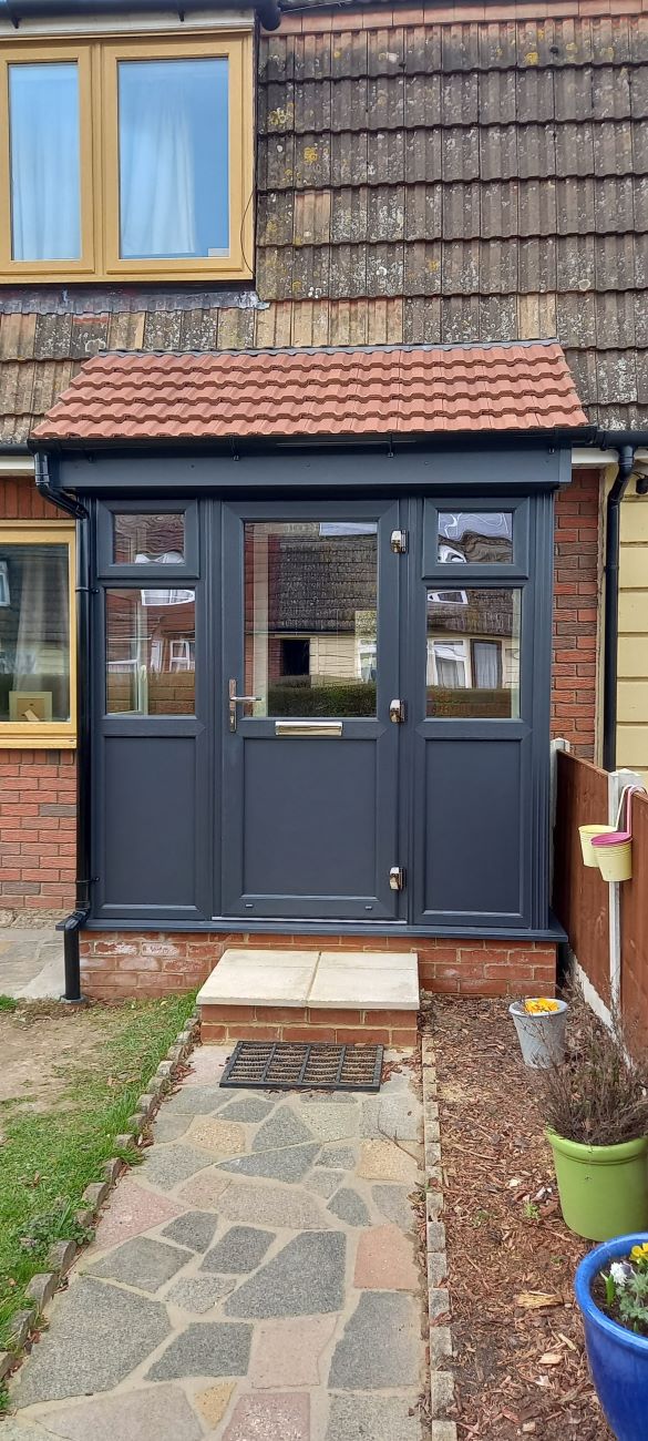 A dark gray porch with glass panels, a red tiled roof, and a stone path leading to it. The house has brown roof tiles, red bricks, and a small garden with a green planter.